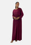 Eine Frau in einem langärmeligen, kastanienbraunen Elara Damen Abaya Maxikleid aus Polyesterstoff steht vor einem schlichten weißen Hintergrund. Mit elegant gefalteten Händen präsentiert sie Ohrringe, Armbänder und offene Sandalen, die ihrem Ensemble einen orientalischen Flair verleihen.