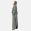Eine gelassene Person präsentiert Elaras fließende hellgraue Abaya und das passende Kopftuch vor einem schlichten Hintergrund. Sie steht im Profil und hat die Hände sanft gefaltet. Das Kleidungsstück fällt elegant um sie herum und verkörpert zeitlose Eleganz.