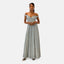 Eine Frau steht selbstbewusst in einem glitzernden, schulterfreien Abendkleid. Das bodenlange Kleid ist silberfarben mit einem eleganten Ausschnitt. Sie trägt metallische Pumps und Ohrringe, ihre Haare sind nach hinten frisiert. Der Hintergrund ist schlicht weiß.