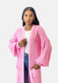 Eine Frau mit langem, dunklem Haar trägt den Elara Oversize Cardigan aus grobem rosa Strick über einem weißen Oberteil und blauen Jeans und steht mit einer Hand in der Tasche vor einem schlichten, hellen Hintergrund für einen lässigen Look und maximalen Komfort.