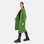Eine Frau steht vor weißem Hintergrund und trägt einen grünen Elara Oversize Cardigan über einem schwarzen Outfit und klobigen Stiefeln. Das Ergebnis ist ein lässiger Look mit maximalem Komfort. Sie blickt in die Kamera und hält eine Seite des Cardigans offen.