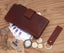 Eine Chunkyrayan Leder-Geldbörse in Braun im Vintage-Look und passende Schlüsselanhänger liegen auf Holz, umgeben von Euro-Scheinen, Münzen, Brille, Taschenuhr und Etikett. Die offene Geldbörse zeigt etwas Bargeld im Inneren.
