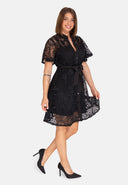 Eine Frau in einem schwarzen kurzärmeligen Spitzenkleid mit Gürtel und schwarzen High Heels steht lächelnd vor einem schlichten weißen Hintergrund.