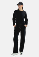 Eine Frau in einem schwarzen Langarmshirt, einer schwarzen Hose mit Kordelzug, weißen Turnschuhen und einer schwarzen Baseballkappe steht mit den Händen in den Taschen da und blickt zur Seite.