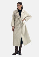 Frau in einem langen, beigen Zweireihermantel mit großen Vordertaschen, schwarzem Unterkleid und braunen kniehohen Stiefeln, steht vor einem schlichten weißen Hintergrund.