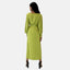 Eine Person mit langem, dunklem Haar steht abgewandt und präsentiert Elaras Eleganz in einem grünen Kleid mit Gürtel und langen Ärmeln sowie schwarzen Stöckelschuhen. Die elegante Schlichtheit des Ensembles wird durch den weißen Hintergrund betont.
