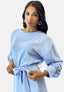 Eine Frau in einem hellblauen Elara-Kleid mit langen Ärmeln und einer Schleife an der Taille steht der Kamera gegenüber. Ihr Look umfasst große Creolen und langes, dunkles Haar. Vor einem weißen Hintergrund mit neutralem Ausdruck strahlt sie Eleganz aus.