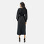 Mit dem Rücken zu ihr trägt eine Person ein elegantes schwarzes Elara-Kleid, das an eine Abaya mit langen Ärmeln erinnert, dazu schwarze High Heels. Der schlichte hellgraue Hintergrund unterstreicht die dezente Eleganz.