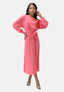 Eine Frau in einem langen rosa Elara-Kleid mit passendem Gürtel posiert selbstbewusst, eine Hand auf der Hüfte. Sie hat lange Haare, große Ohrringe und Riemchensandalen mit hohen Absätzen. Der schlichte weiße Hintergrund unterstreicht ihre anmutige Präsenz.