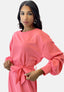 Eine Frau in einem rosa Elara-Kleid mit langen Ärmeln und gebundener Taille steht elegant vor einem neutralen Hintergrund und trägt große Creolen. Ihr langes Haar fällt herab und verkörpert zeitlose Eleganz.