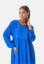 Eine Frau in einem blauen, langärmeligen Elara Damen Abaya Maxikleid steht anmutig da, eine Hand in der Tasche, die andere am Kinn. Ihr Pferdeschwanz, die goldenen Ohrringe und das Armband verleihen ihr Eleganz vor dem schlichten weißen Hintergrund, der den Polyesterstoff hervorhebt.