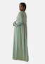 Eine Frau in einem Elara Damen Abaya Maxikleid, das orientalisches Flair verströmt, steht vor einem schlichten Hintergrund. Ihr langes Haar ist zurückgebunden, und ihre goldenen Ohrringe ergänzen den fließenden Polyesterstoff ihres hellgrünen Kleides perfekt, während sie über ihre Schulter blickt.