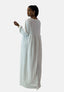 Eine Frau in einem weißen, langärmeligen Elara Damen Abaya Maxikleid blickt zur Seite. Ihr Haar ist zu einem Pferdeschwanz mit orientalischer Raffinesse frisiert, ergänzt durch goldene Ohrringe und Armbänder. Der Hintergrund ist schlicht weiß.