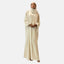Eine Person in einer hellbeigen Elara-Abaya und dem dazu passenden Hijab verkörpert zeitlose Eleganz und steht anmutig vor einem schlichten Hintergrund. Die Abaya hat weite Ärmel und ein fließendes Design. Mit sanft gefalteten Händen wird der Look durch schlichte Sandalen abgerundet.