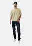 Ein Mann mit Brille, einem beigen T-Shirt von Elara, dunklen Jeans und weißen Turnschuhen steht vor einem schlichten weißen Hintergrund, blickt leicht nach oben und hat eine Hand in der Tasche.