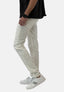 Eine Person in cremefarbenen Elara-Jeans, weißen Elara-Sneakers mit schwarzen Streifen, einem schwarzen Elara-T-Shirt und einer schwarzen Elara-Armbanduhr steht vor einem schlichten weißen Hintergrund.
