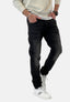 Ein Mann in einem hellen Sweatshirt, schwarzen Jeans von Elara im Waschlook und weißen Sneakers mit schwarzen Streifen steht mit den Händen in den Taschen vor einem schlichten Hintergrund.
