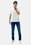 Ein Mann mit Brille, einem weißen T-Shirt von Elara, Bluejeans im Waschlook und weißen Turnschuhen steht nachdenklich vor einem schlichten hellen Hintergrund und berührt sein Kinn.