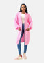 Eine Frau blickt nach vorne und trägt einen Elara Oversize Cardigan in Pink über einem weißen Top und Blue Jeans für einen lässigen Look. Sie trägt pink-weiße Sneakers, Creolen, lange Haare und hat ihre Hände für maximalen Komfort in den Taschen des Cardigans vergraben.
