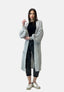 Eine Frau steht vor einem hellen Hintergrund in einer übergroßen Elara-Strickjacke, einem schwarzen Top, einer Hose mit Umschlag und weißen Sneakers. Glattes braunes Haar, ein neutraler Gesichtsausdruck und eine erhobene Hand runden ihren Look ab.