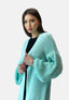 Eine Frau mit glattem braunem Haar trägt einen Elara Oversize Cardigan in Hellblau über einem schwarzen Oberteil, steht vor einem schlichten weißen Hintergrund und blickt mit neutralem Gesichtsausdruck leicht zur Seite.