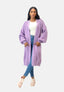 Eine Frau steht vor einem hellen Hintergrund und trägt einen Elara Oversize Cardigan aus lavendelfarbenem Strick mit Fronttaschen, ein weißes Top, blaue Jeans und rosa Sneakers. Ihr lässiger Look und die Hände in den Taschen versprechen maximalen Komfort.