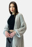 Eine Frau mit glattem braunem Haar trägt ein schwarzes geripptes Crop-Top, schwarze Jeans mit hoher Taille und einen Elara Oversize Cardigan in Hellgrau. Sie steht vor einem schlichten Hintergrund und strahlt einen lässigen Look und Selbstvertrauen aus.